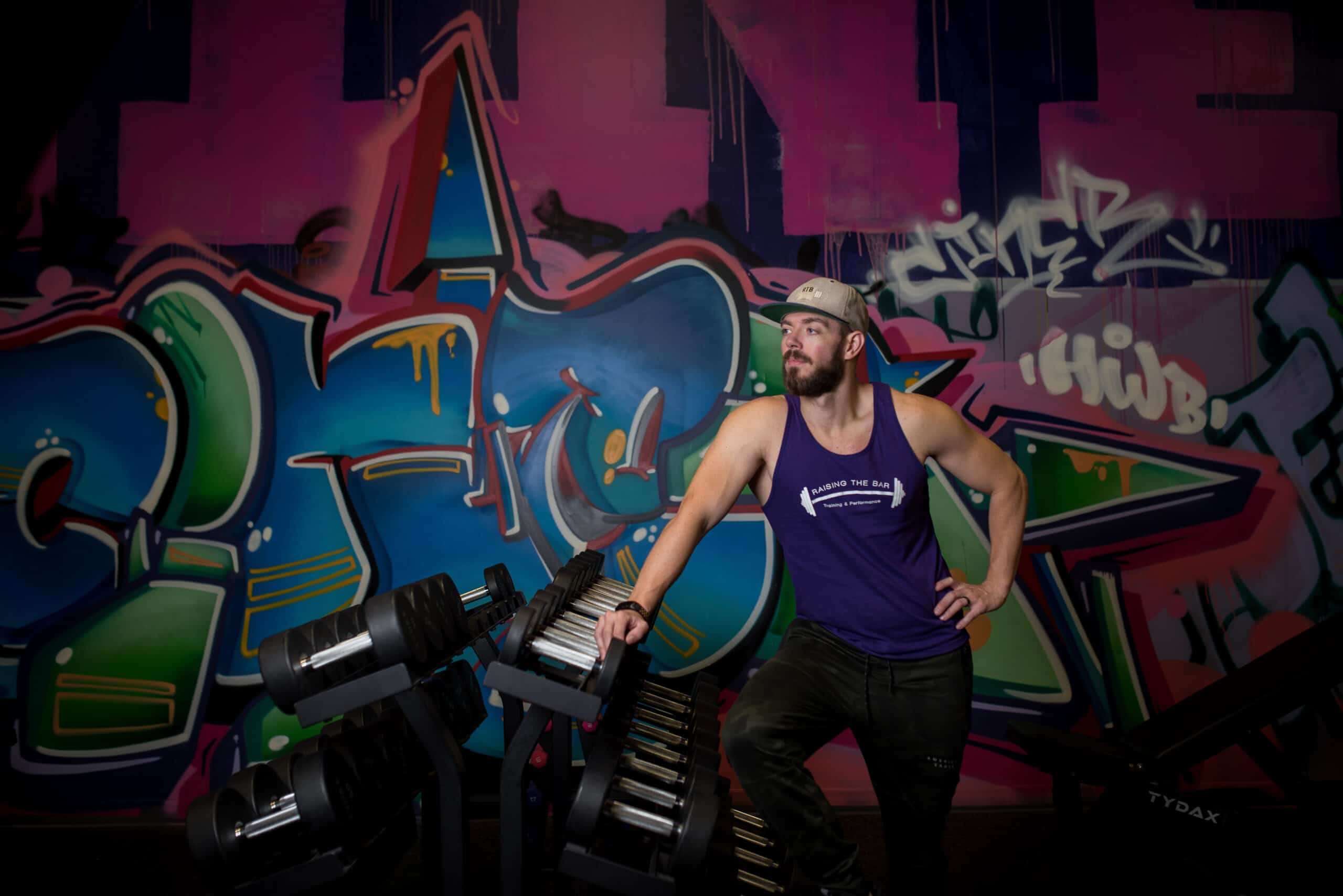 The owner of Raising The Bar Training Shaun Smears standing in front of a decorative graphiti wall of his gym.